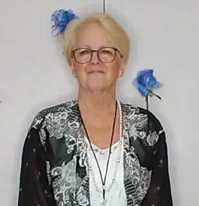 A woman with blonde hair and glasses standing in front of blue flowers.